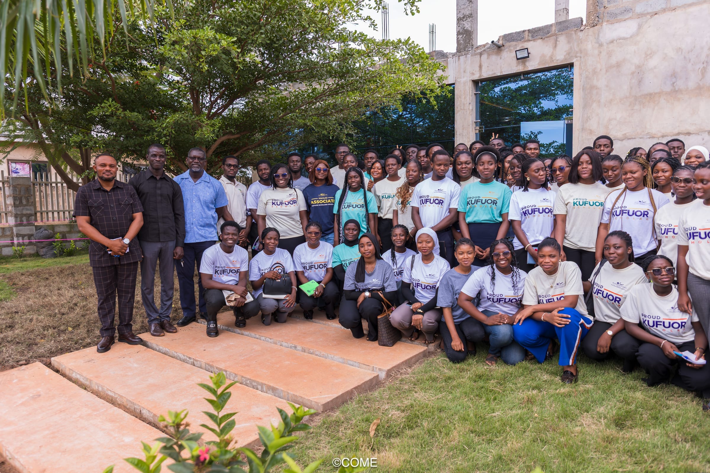 Large outdoor group of young adults and mentors on a lawn - many in program shirts including Proud Kufuor Scholar - showing cohort scale and community.